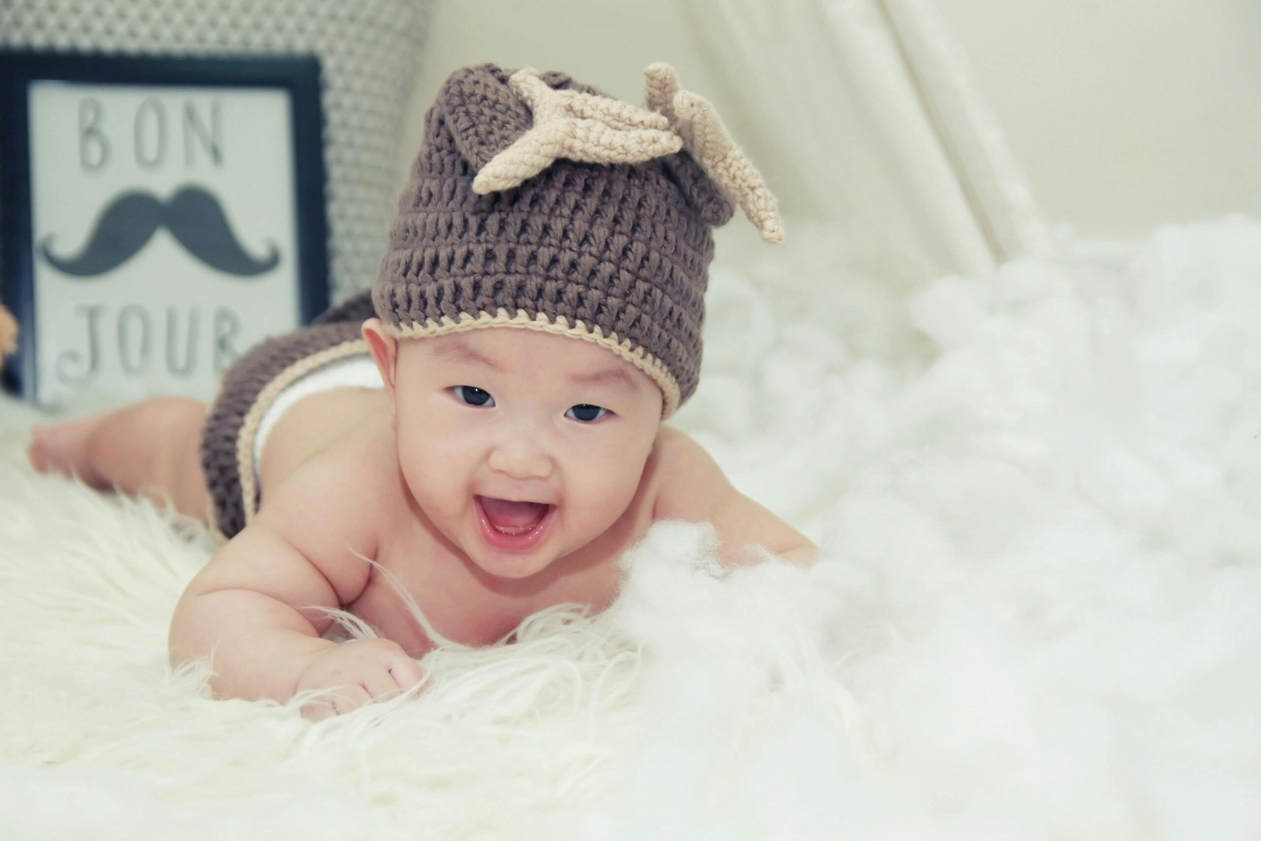 Baby Face Predictor – AI Baby Generator 5 A joyful baby wearing a brown knitted hat lies on a soft surface, radiating happiness.